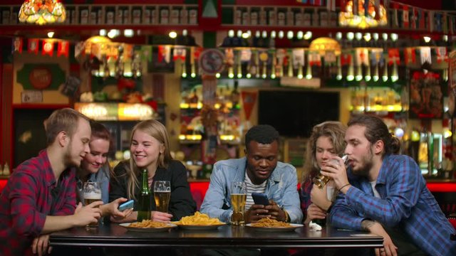 Cheerful students sitting at a table in a bar drinking beer, eating chips and watching photos on a smartphone screen discussing photos