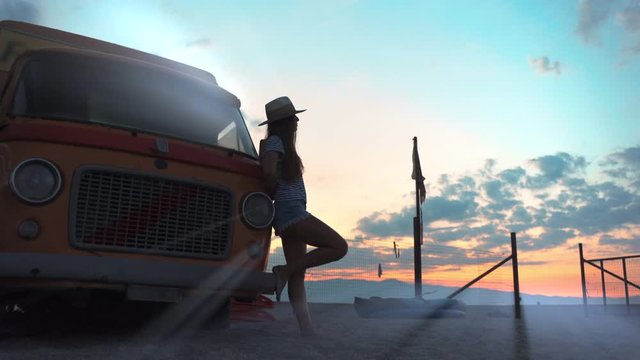 Thinking girl in straw hat leaning on amazing red and orange retro car and looking in distance. Dreaming tourist female having rest at the sea beach. Beautiful retro style girl silhouette in