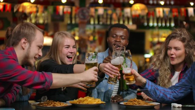 Close-up plan a company of friends makes a toast and everybody knocks, check their glasses with beer, drink, and laugh. Students meeting in a bar, restaurant