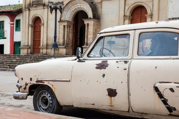 Antique rusty car parked next to the Parish Church of the small town of Ventaquemada in Colombia