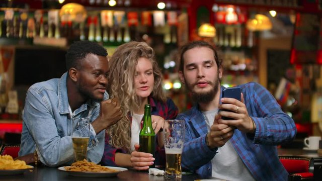 Two men and a woman look at the phone screen while sitting in a bar and prepare to take a shared selfie. Cheerful youth company at the bar