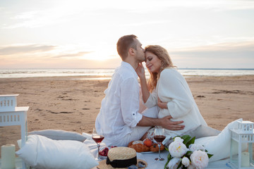 Picnic at sunset. Man and a pregnant woman sit on a bedspread, on the beach, hugging, kissing against low tide.
