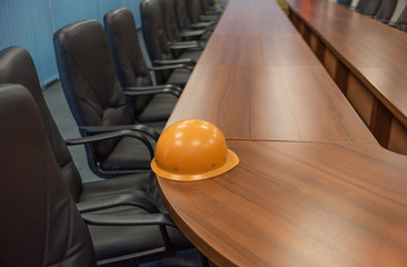 An orange safety helmet left on a long table in a discussion room, with a row of black leather chairs stretching from foreground to background.