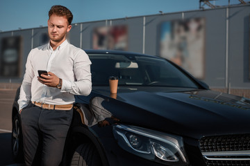 Young successful businessman in a white shirt checking smartphone standing near his luxary car. Disposable cup of hot coffee on a motor hood.