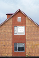 Part of a classic two-story red brick house against a clear sky. Triangular roof with a pipe and windows. Copy space for articles on affordable housing under the state program or mortgage lending.