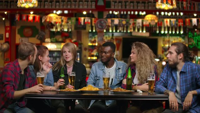 A multi-ethnic group of young men and women drink beer at a bar and eat chips and cheerfully debate about the university. Laughing at a joke