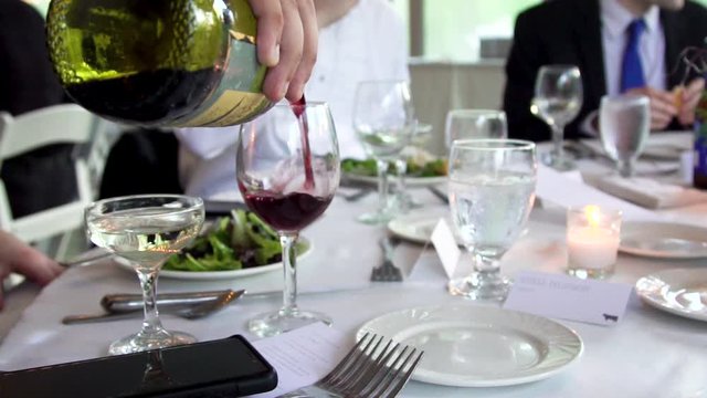 slow motion footage of a man pouring red wine in a glass during a wedding events dinner. the elegant plate setting and food is very luxurious at the guest await their meal in celebration. 