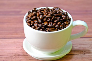 White coffee cup filled up with roasted coffee beans placed on wooden table.