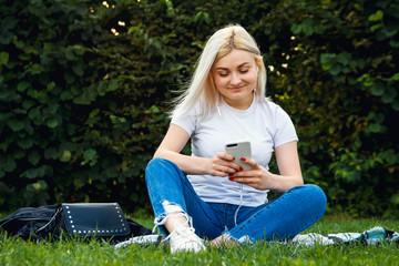 Beautiful blonde girl in headphones and smartphone in hands sits on green grass. She is talking on the phone through headphones. Lifestyle concept. Copy, empty space for text