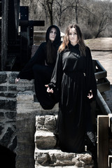 Young Women with dark long hair in black robes in front of an Old Wooden Water Mill. Witches. Halloween concept. Witchcraft and Magic. 