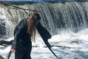Young  tall Woman with dark long hair in black robes in front of the Waterfall. Witches. Halloween concept. Witchcraft and magic. 