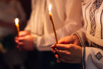 newlyweds in embroidered clothes hold lighted candle in church.