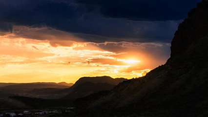 Obraz premium The setting sun casts an orange glow over the mountain landscape as dark storm clouds fill the sky
