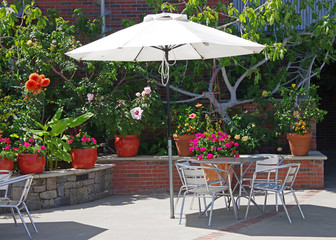 Coffee house style outdoor chairs and table under an umbrella in a lush garden setting on a bright...