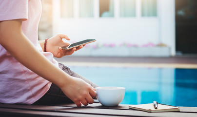 Young asian woman resting at the pool side she using smartphone online to work and hold coffee to...