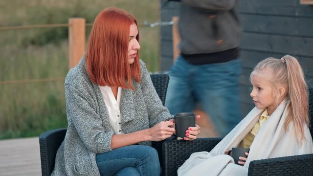 A young Caucasian family couple with a baby daughter sits on the terrace in chairs and drinks hot drinks. Against the background of a small modern rustic wooden house. Father is cooking barbecue. Summ