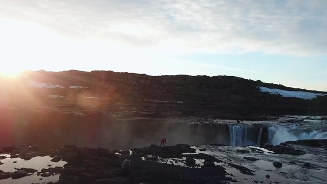 Aerial, drone shot, around a woman, standing on the top of the Godafoss falls, taking pictures of the waterfall of the gods, at the Skjalfandafljot river, at sunset, on a sunny evening, in Iceland