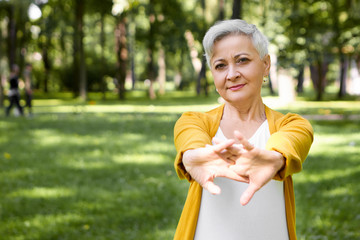 Outdoor image of attractive gray haired retired woman stretching her arms muscles, warming up body before morning run in park. People, sports, healthy, fitness, aging, recreation and activity concept