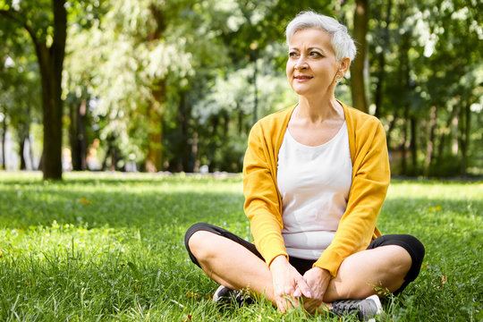 Mature people, age, retirement and wellness concept. Summer image of good looking athletic female pensioner with pixie hairstyle sitting on grass with legs crossed, admiring beautiful sunny day