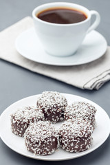 Swedish sweets chocolate balls or chokladbollar, made from oats, cocoa, butter and coconut, on a white plate, served with coffee, vertical