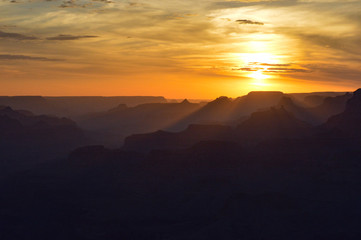 Sunset at the Grand Canyon as the sun is about to slip over the horizon, leaving visible rays between the peaks and ridges, beautifully-colored sky, gradient mountain ranges
