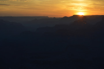 Sunset at the Grand Canyon as the sun is about to slip over the horizon, leaving a small sun flare between the peaks and ridges, beautifully-colored sky, gradient mountain ranges