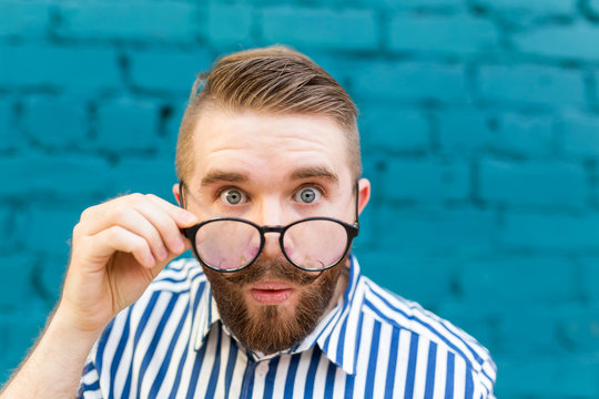 Close-up portrait of a curious surprised young man in glasses with a mustache and beard posing on a background of blue blurred brick wall. Concept of surprise and shocking information.