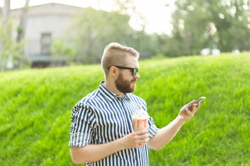 Naklejka na ściany i meble Side view of a stylish young man with a beard holding a milkshake and admiring the city views walking in the park on a warm summer day. The concept of rest and relaxation.