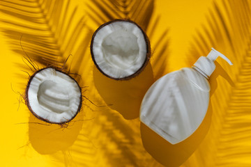 Minimalistic beauty still life. Two halves of chopped coconut and white bottle of cream with shadows from palm leaves on yellow background. Creative fashion concept. Top view.