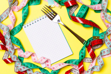 Top view of notebook with fork surrounded with colorful measuring tapes on yellow background. Copy space with diet planning