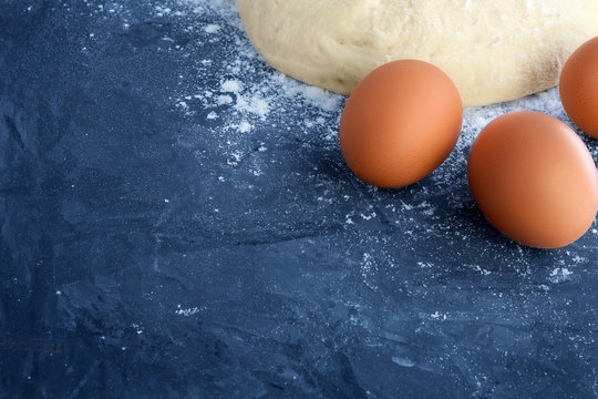 A ball of raw bread dough, sprinkled white wheat flour and three brown eggs in the corner of the picture on a blue textured concrete background with empty spaces for text and other design elements