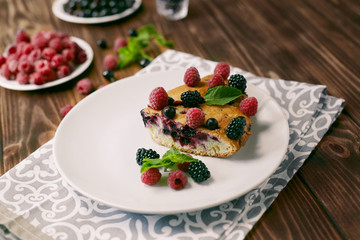 Piece of pie with blueberries, rasberry and mint for dessert on a white plate, napkin on a wooden boards background
