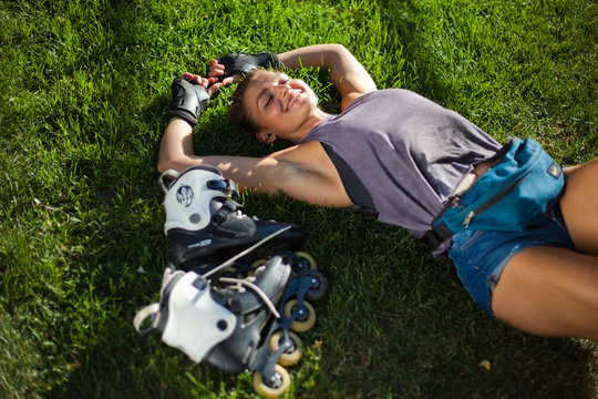 Young cheerful sport woman roller skater resting lying on the lawn grass in the park and listens to music with headphones