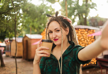 Attractive woman with an informal haircut dreadlocks makes selfie portrait and holding cup of coffee on background of garlands in the evening