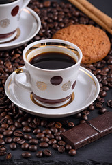 Fototapeta premium white coffee mug on a saucer on a stone table with scattered coffee beans, oatmeal cookies, chocolate and cinnamon stick