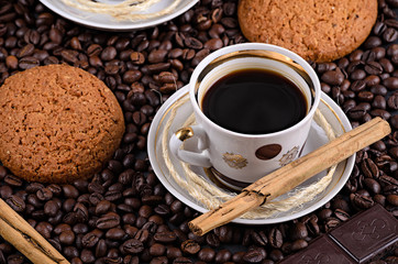 white coffee mug on a saucer on a stone table with scattered coffee beans, oatmeal cookies, chocolate and cinnamon stick