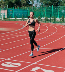 Young cheerful woman runner in sportswear running on stadium track with red coating outdoors
