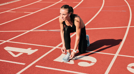 Fototapeta na wymiar Young sprinter woman in sportswear tying shoelaces before a race on a red-coated stadium track at sunny bright day