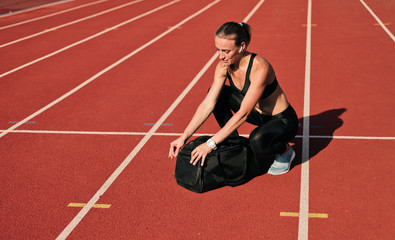 Young woman athlete in sportwear getting ready for workout and opening sports bag on the track of stadium at bright sunny day
