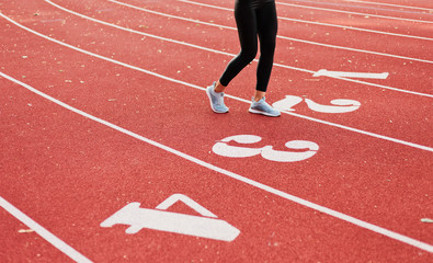 Young runner woman in sportwear run on stadium red track with numbers. Crop photo of female legs in leggings and sneakers