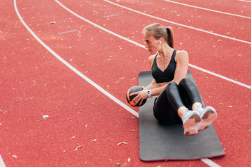 Young fit woman perfoms her body while doing exercise with medicine ball on mat at stadium track with red coating. Healthy lifestyle