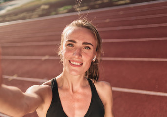 Young attractive woman athlete in sport clothes taking selfie on stadium track with red coating on...