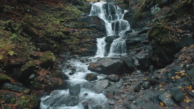 pictorial foaming waterfall over large wet stones surrounded by green moss and brown autumn leaves aerial view. Shipot waterfall, Carpathian mountains. 4K
