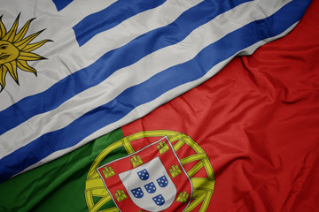waving colorful flag of portugal and national flag of uruguay.