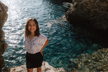 Adventurous young latin woman looking sideways on the sea rocks. Woman on cliff, with the sunset. Jovial near the sea on vacation in the Mediterranean with turquoise blue water.