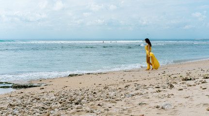 Obraz premium Woman in long yellow dress on rocky beach on Bali, Indonesia. Summer time. Dark hair brunette