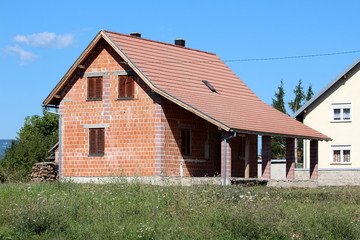 Fototapeta na wymiar New unfinished small red brick family house with front porch surrounded with uncut grass and other houses on warm sunny summer day