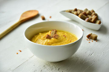 Yellow zucchini and carrot puree soup in a white bowl. White wooden background. Croutons.