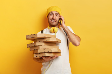 Photo of pizza man receives orders from customers via smart phone, holds many carton boxes with fast food, has unpleasant look to speak with dissatisfied client. Service and delivering concept
