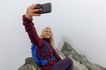 Young Adventurous Girl on top of a rugged rocky mountain taking a selfie during a cloudy summer...
